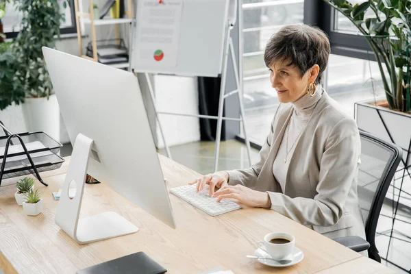 Lächelnde und stilvolle Geschäftsfrau mittleren Alters, die in moderner Büroumgebung am Computer arbeitet — Stockfoto