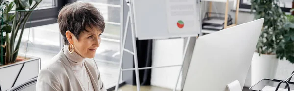 Positiva donna d'affari di mezza età che guarda il monitor del computer mentre lavora in ufficio, banner — Foto stock