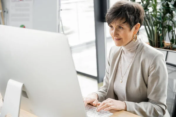 Empresaria optimista de mediana edad escribiendo en el ordenador mientras trabaja en la oficina contemporánea - foto de stock