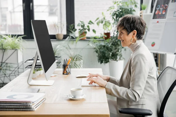 Вид збоку бізнес-леді середнього віку, що працює на комп'ютері біля чашки кави та документів в офісі — стокове фото