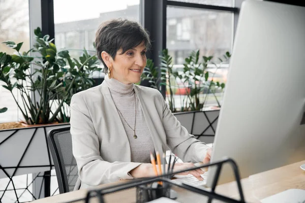 Successful middle aged entrepreneur smiling and working on computer in modern office — Stock Photo