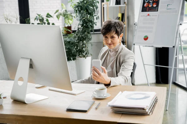 Счастливая зрелая деловая женщина смотрит на смартфон рядом с компьютером, ноутбуки и документы на рабочем месте — стоковое фото