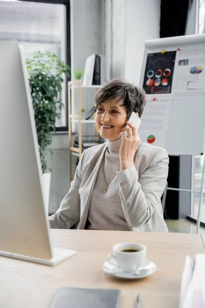 Joyful middle aged businesswoman talking on smartphone near coffee cup in modern office — Stock Photo