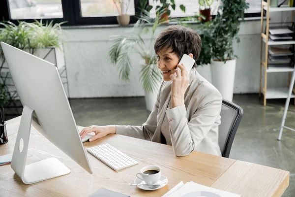 Sonriente gerente de mediana edad hablando en el teléfono móvil cerca de la computadora portátil y café en la oficina contemporánea - foto de stock