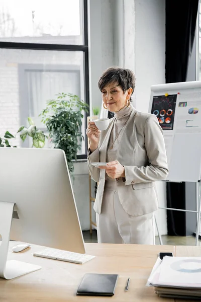Улыбающаяся деловая женщина средних лет, стоящая с чашкой кофе и глядя на монитор компьютера — стоковое фото