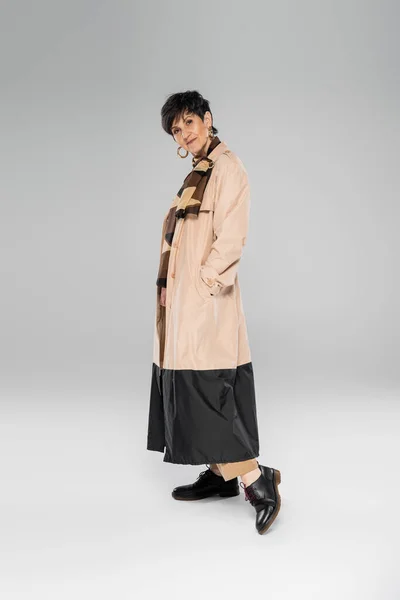 Femme d'âge moyen en tenue d'automne tendance regardant la caméra sur gris, trench coat, écharpe, pleine longueur — Photo de stock