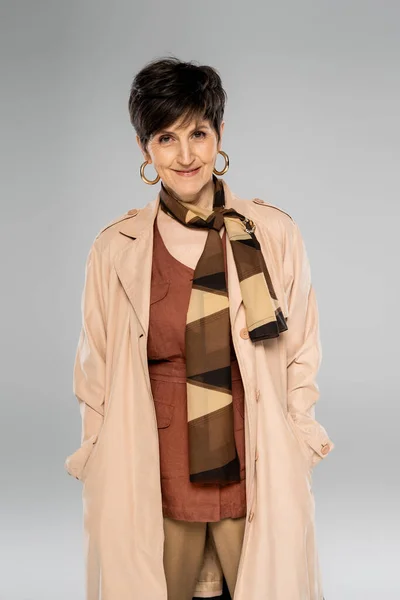 Mujer madura feliz mirando a la cámara en gris, moda de otoño, gabardina, bufanda, pendientes de aro - foto de stock