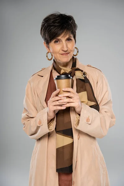 Donna matura alla moda con caffè per andare sul grigio, stile autunno, trench, sciarpa, orecchini a cerchio — Foto stock