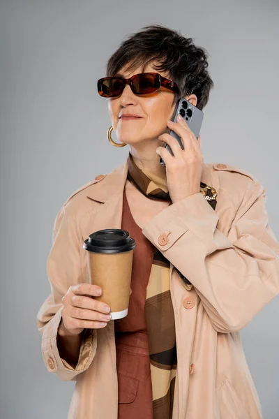 Trendy middle aged businesswoman in sunglasses with paper cup, talking on smartphone on grey — Stock Photo