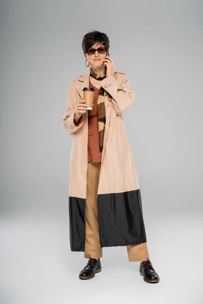 Elegante mujer de negocios de mediana edad en gafas de sol, con café para llevar, hablando en el teléfono inteligente en gris - foto de stock