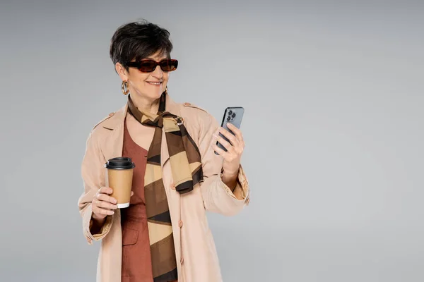 Happy middle aged businesswoman with smartphone and paper cup on grey, autumn fashion — Stock Photo