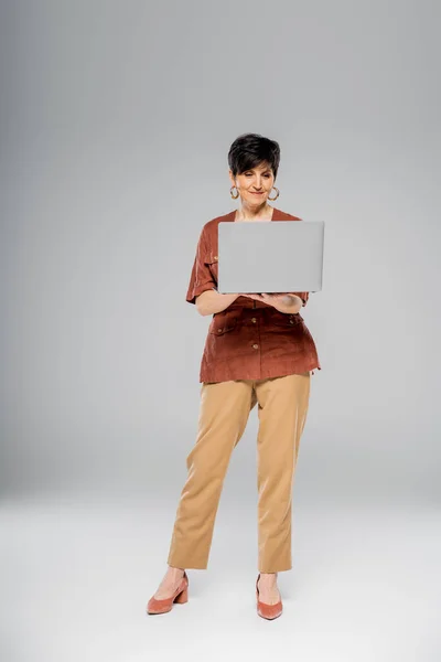 Mature businesswoman in brown jacket using laptop on grey, autumn, seasonal attire, fashion trend — Stock Photo