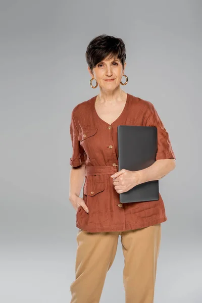 Élégante femme d'affaires d'âge moyen avec ordinateur portable et la main dans la poche sur gris, veste marron, mode automne — Photo de stock