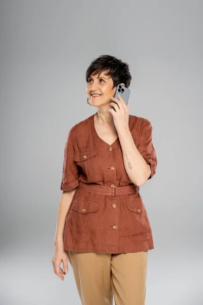 Joyful mature businesswoman talking on smartphone on grey, autumn style, fashion-forward — Stock Photo
