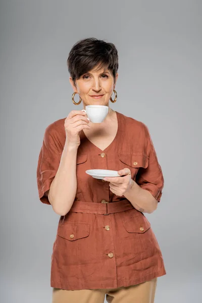 Morena mujer de mediana edad en chaqueta de lino marrón bebiendo café en gris, moda de otoño - foto de stock