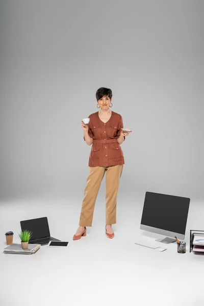 Maduro mujer de negocios en ropa de otoño con estilo, taza de café, dispositivos digitales, documentos en gris - foto de stock