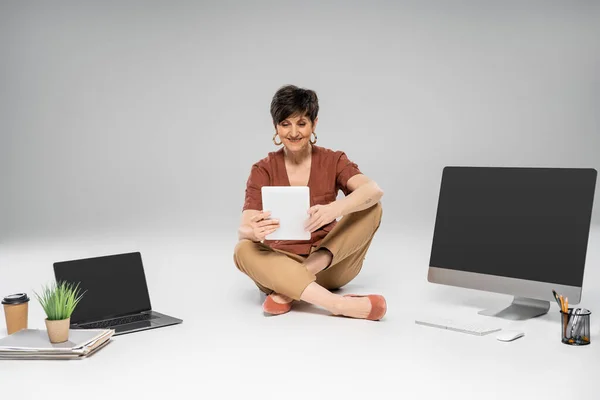 Joyful middle aged businesswoman with digital tablet sitting near devices and documents on grey — Stock Photo