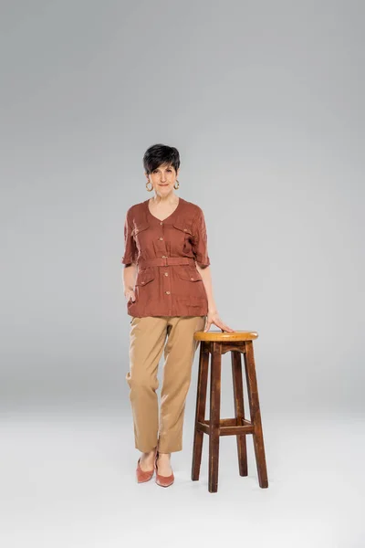 Осенняя мода, женщина средних лет в модной одежде рядом с высоким стулом на сером, полная длина — стоковое фото