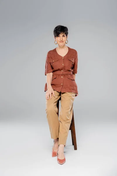 Automne mode, femme d'âge moyen en tenue tendance assise sur tabouret haut sur gris, pleine longueur — Photo de stock