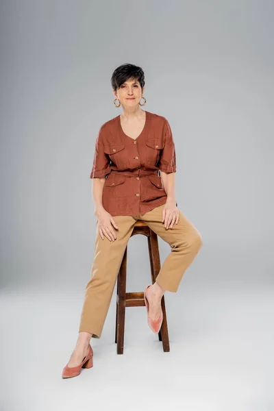 Осенняя мода, зрелая брюнетка в стильной одежде, сидящая на высоком стуле на сером, полной длины — стоковое фото