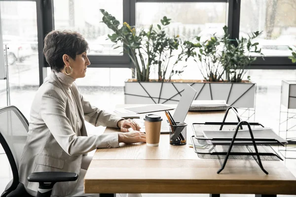 Seitenansicht einer selbstbewussten Geschäftsfrau mittleren Alters, die in der Nähe eines Imbisses im modernen Büro am Laptop arbeitet — Stockfoto