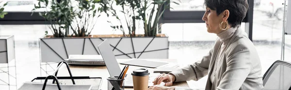 Вид сбоку опытного менеджера среднего возраста, работающего на ноутбуке рядом с кофе, чтобы пойти в офис, баннер — стоковое фото