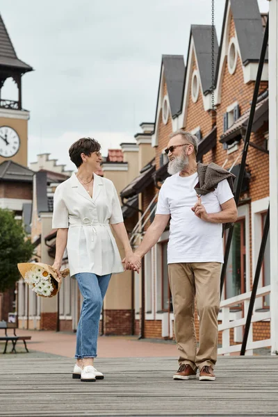 Счастливая пожилая пара в солнечных очках, улыбающаяся и держащаяся за руки, идущая вместе, по магазинам, летом — стоковое фото