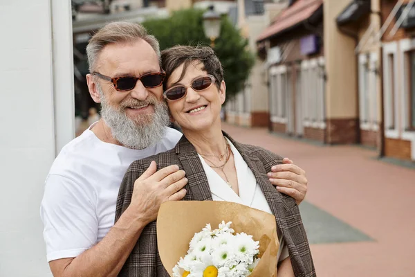 Счастливая пожилая пара в солнечных очках улыбается и смотрит в камеру, летом, пожилой мужчина обнимает женщину — стоковое фото