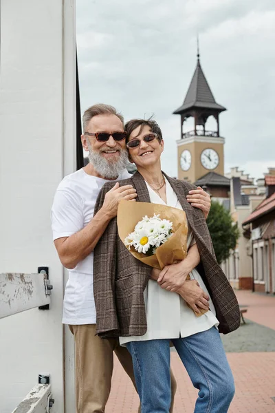 Счастливая пожилая пара в солнечных очках, мужчина обнимает женщину цветами, лето, розетка, букет, романтика — стоковое фото