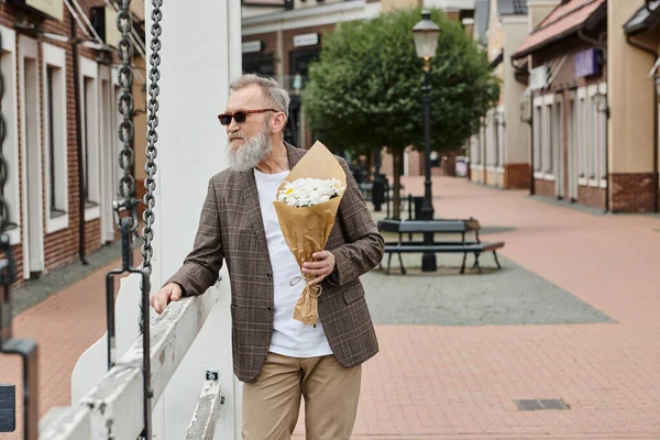 Старший мужчина с бородой и солнцезащитными очками в руках букет цветов, городской фон, стильный наряд — стоковое фото