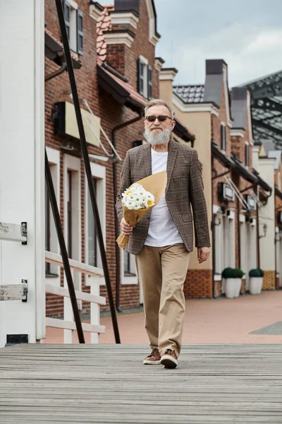 Senior man with beard and sunglasses holding bouquet of flowers, walking on urban street, stylish — Stock Photo