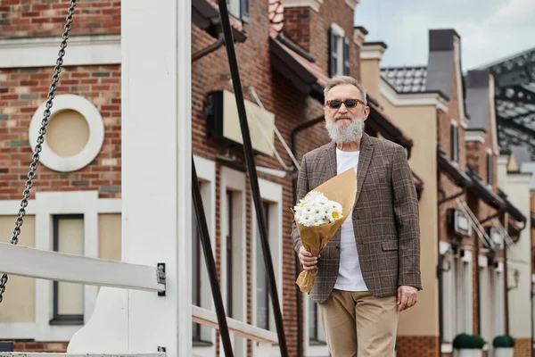 Senior man with beard and sunglasses holding bouquet of flowers, standing on urban street, stylish — Stock Photo