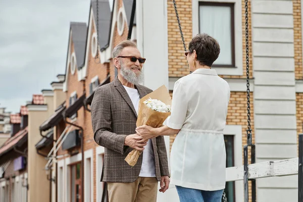Щасливий літній чоловік з бородою і модними сонцезахисними окулярами дарує букет жінці на вулиці, дата, романтика — стокове фото