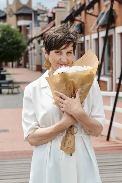 Mujer anciana sosteniendo ramo de flores y mirando a la cámara, telón de fondo urbano, verano, traje blanco - foto de stock