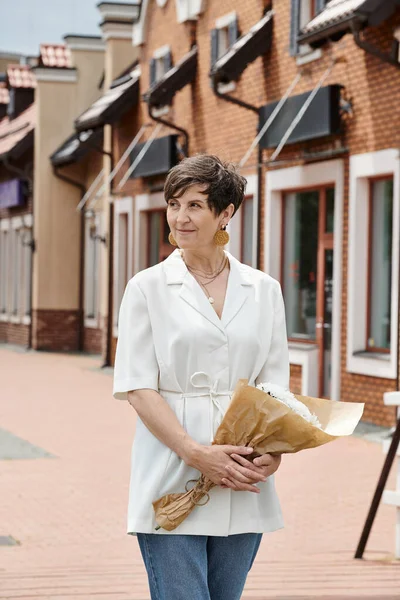 Femme âgée tenant bouquet de fleurs et regardant loin sur fond urbain, été, tenue blanche — Photo de stock