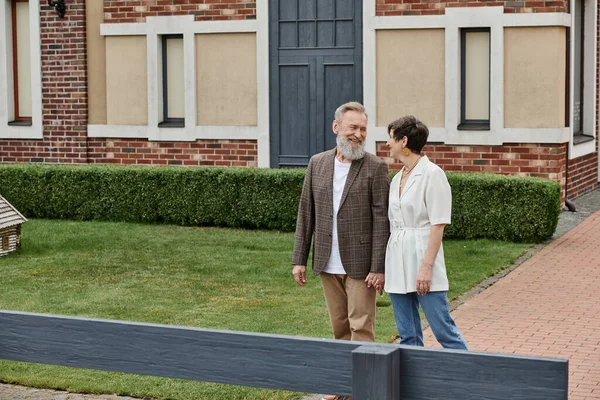 Cheerful elderly couple, man and woman standing near house and looking at each other, love, romance — Stock Photo