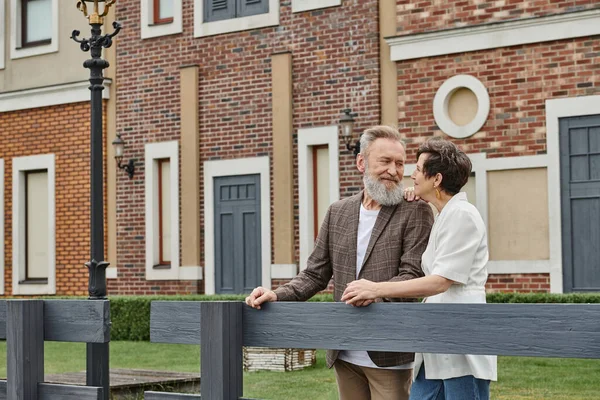 Щаслива старша пара, чоловік і жінка стоять біля паркану поруч з будинком, дивлячись один на одного, романтика — стокове фото