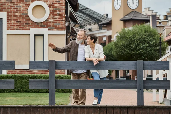 Joyful elderly couple, man pointing away near woman, togetherness, urban backdrop, aging population — Stock Photo