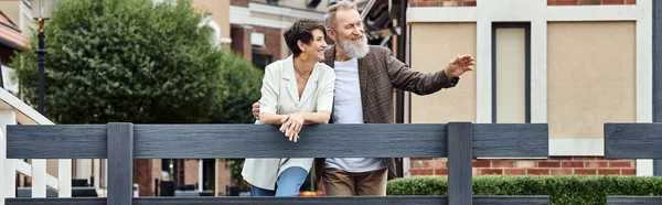 Alegre casal idoso, homem apontando para longe, união, pano de fundo urbano, envelhecimento da população, bandeira — Fotografia de Stock