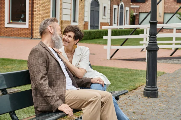 Feliz anciana mirando al hombre barbudo, romance, marido y mujer sentados en el banco, urbano - foto de stock