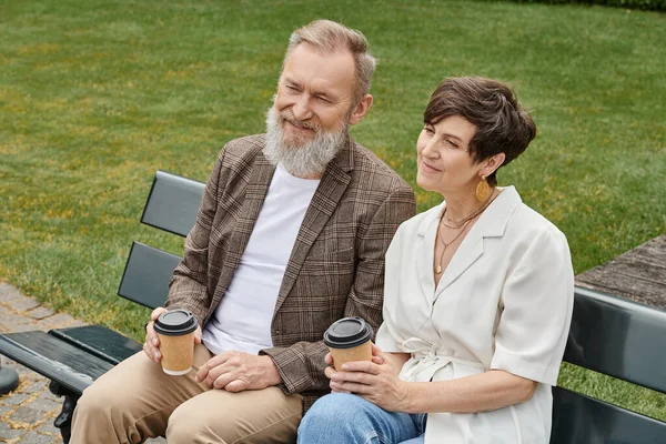 Feliz anciano y mujer sentado en el banco, sosteniendo tazas de papel con café, pareja de ancianos, romance - foto de stock