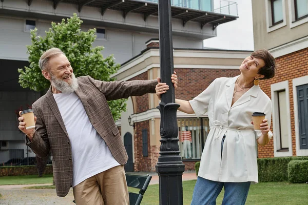 Счастливая пожилая пара проведение кофе идти, стоя рядом уличный фонарь, мужчина и женщина, пожилые люди, романтика — стоковое фото