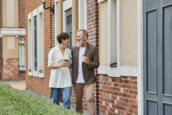 Happy senior couple holding coffee to go, walking near building, elderly man and woman, laughter — Stock Photo