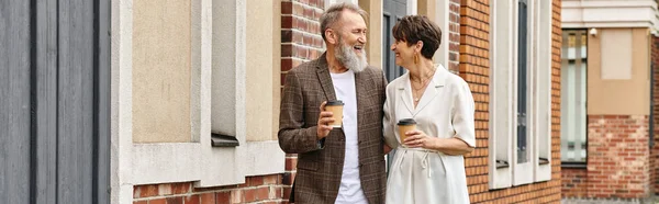 Feliz pareja de ancianos caminando con café cerca del edificio, anciano hombre y mujer, risa, pancarta - foto de stock