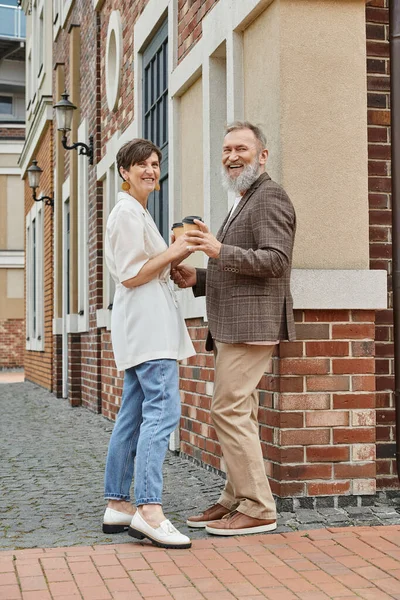 Счастливая пожилая пара держит кофе, чтобы пойти, здание, пожилой мужчина и женщина смотрят в камеру, свидание — стоковое фото