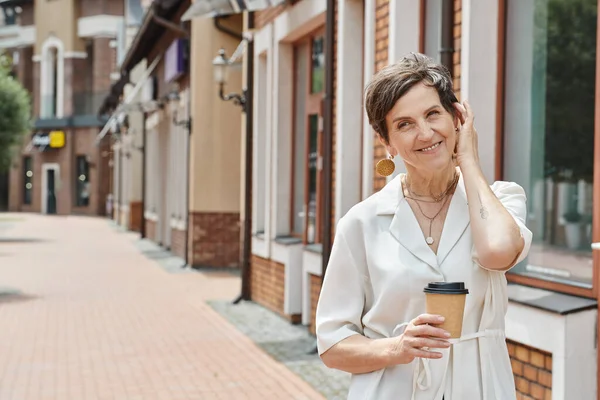 Heureuse femme âgée tenant tasse en papier avec café à emporter, ajustement des cheveux courts, toile de fond urbaine, sourire — Photo de stock