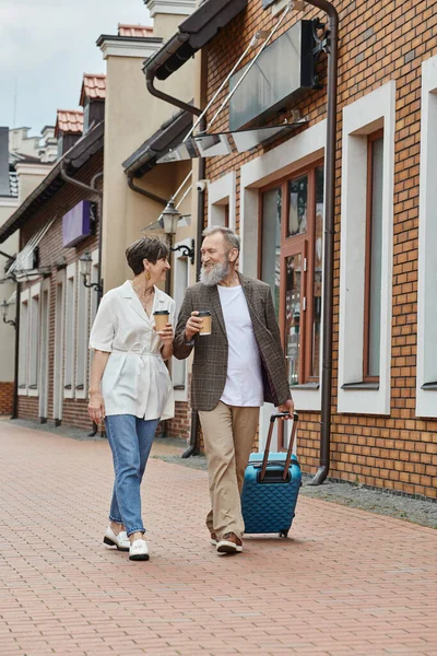 Feliz pareja de ancianos, hombre y mujer caminando con café para llevar y equipaje en la calle, estilo de vida urbano - foto de stock