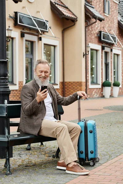Hombre mayor alegre usando el teléfono inteligente y sentado en el banco cerca del equipaje, usando gadget, edad en tecnología - foto de stock