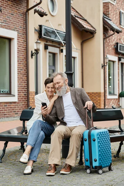 Пожилая пара, счастливый бородатый мужчина держит смартфон, сидит с женщиной на скамейке, багаж, гаджет — стоковое фото
