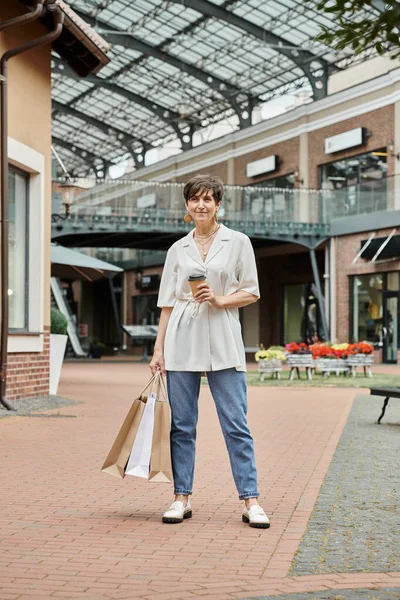 Seniorin mit kurzen Haaren hält Einkaufstüten und Kaffee in der Nähe des Outlets im Freien, ältere — Stockfoto